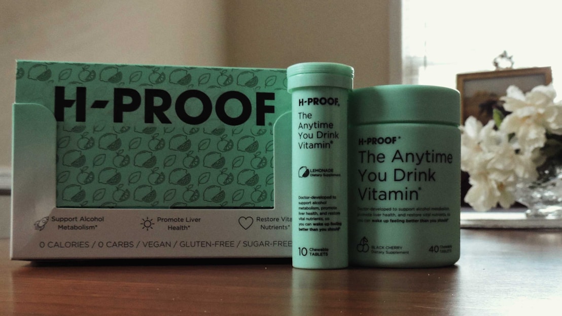 A collection of H-PROOF products including a box and two containers labeled as 'The Anytime You Drink Vitamin.' The packaging highlights health benefits such as supporting alcohol metabolism and promoting liver health. The products are vegan, gluten-free, and sugar-free. They are placed on a wooden surface with a vase of white flowers in the background.