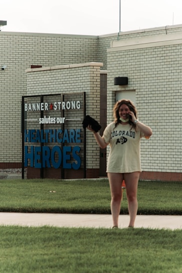 A person wearing a Colorado t-shirt and talking on the phone stands in front of a building with a sign reading 'Banner Strong salutes our Healthcare Heroes'. The building is made of light-colored bricks and the person appears happy.