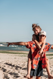 A young couple enjoys a playful moment on a sandy beach. One person is giving the other a piggyback ride, with the person on top smiling and pointing towards something in the distance. The sky is clear, and the ocean is visible in the background.
