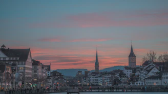 A historic city skyline at dusk, with softly glowing lights reflecting on calm river waters.
