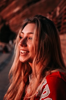 A person with long blonde hair smiles warmly, wearing a red jacket adorned with white lettering. The image is captured in warm, glowing light, highlighting the person's joyful expression.