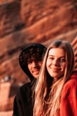 A young man and woman are seated closely together, smiling warmly in a setting with warm, earthy tones in the background. Both have long hair, and the woman is wearing a red hoodie, while the man is in a black one.