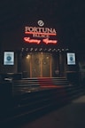 A neon-lit casino entrance with a red carpet leading up to large double doors. The sign reads 'Fortuna Palace Luxury Games' in bright red and white neon lights. The dimly lit atmosphere suggests a night-time setting. There are two digital displays on either side of the entrance showing promotional content.