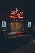 A neon-lit casino entrance with a red carpet leading up to large double doors. The sign reads 'Fortuna Palace Luxury Games' in bright red and white neon lights. The dimly lit atmosphere suggests a night-time setting. There are two digital displays on either side of the entrance showing promotional content.