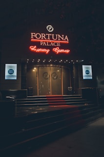A neon-lit casino entrance with a red carpet leading up to large double doors. The sign reads 'Fortuna Palace Luxury Games' in bright red and white neon lights. The dimly lit atmosphere suggests a night-time setting. There are two digital displays on either side of the entrance showing promotional content.