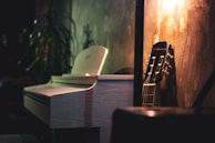 A dimly lit room features a white piano with a sleek, modern design positioned next to a wooden acoustic guitar leaning against a textured wall. The warm glow from a nearby light casts soft shadows, creating an inviting and serene atmosphere. In the background, a houseplant adds a touch of nature.