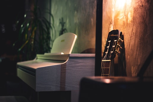 A dimly lit room features a white piano with a sleek, modern design positioned next to a wooden acoustic guitar leaning against a textured wall. The warm glow from a nearby light casts soft shadows, creating an inviting and serene atmosphere. In the background, a houseplant adds a touch of nature.