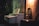 A dimly lit room features a white piano with a sleek, modern design positioned next to a wooden acoustic guitar leaning against a textured wall. The warm glow from a nearby light casts soft shadows, creating an inviting and serene atmosphere. In the background, a houseplant adds a touch of nature.