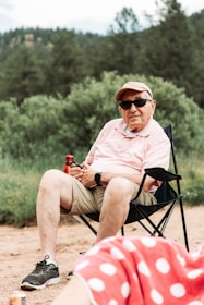 An elderly person wearing a pink shirt and cap, seated on a folding chair in an outdoor setting. The individual is holding a drink bottle and wearing sunglasses, with a watch on their wrist. The background is composed of lush greenery and trees. In the foreground, a person in a red polka-dot fabric is partially visible.