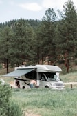 A motorhome is parked in a lush green campsite surrounded by tall pine trees. Two people are engaged in activities near the vehicle, one wearing a shirt and the other shirtless. The motorhome has an extended awning providing shade.