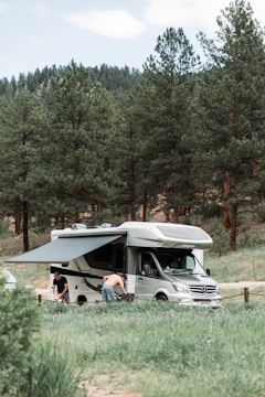 A motorhome is parked in a lush green campsite surrounded by tall pine trees. Two people are engaged in activities near the vehicle, one wearing a shirt and the other shirtless. The motorhome has an extended awning providing shade.