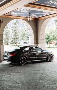 A sleek black car is parked under an ornate ceiling with blue and gold accents. The setting appears to be a covered entryway or drive-through with large stone arches and decorative ironwork. Trees and a lamppost can be seen in the background, suggesting an elegant or upscale environment.