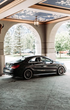 A sleek black car is parked under an ornate ceiling with blue and gold accents. The setting appears to be a covered entryway or drive-through with large stone arches and decorative ironwork. Trees and a lamppost can be seen in the background, suggesting an elegant or upscale environment.