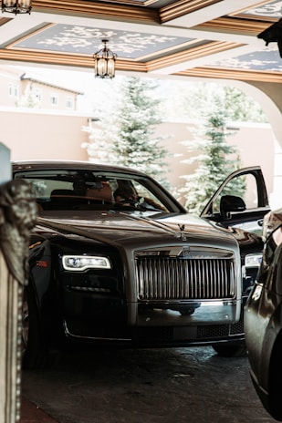 A luxurious car with a sleek black exterior is parked in a covered area with decorative ceiling designs. The vehicle's front grille is prominently displayed, and one of the doors is slightly open. Trees and a building are visible in the background.