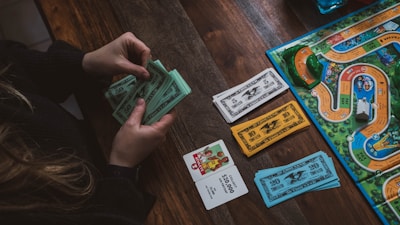 A person is holding and counting green and blue play money bills at a wooden table. On the table, there is also a board game with colorful paths, miniature cars, card decks, and more play money in white and orange colors.