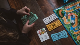 A person is holding and counting green and blue play money bills at a wooden table. On the table, there is also a board game with colorful paths, miniature cars, card decks, and more play money in white and orange colors.