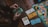 A person is holding and counting green and blue play money bills at a wooden table. On the table, there is also a board game with colorful paths, miniature cars, card decks, and more play money in white and orange colors.