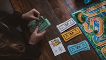 A person is holding and counting green and blue play money bills at a wooden table. On the table, there is also a board game with colorful paths, miniature cars, card decks, and more play money in white and orange colors.