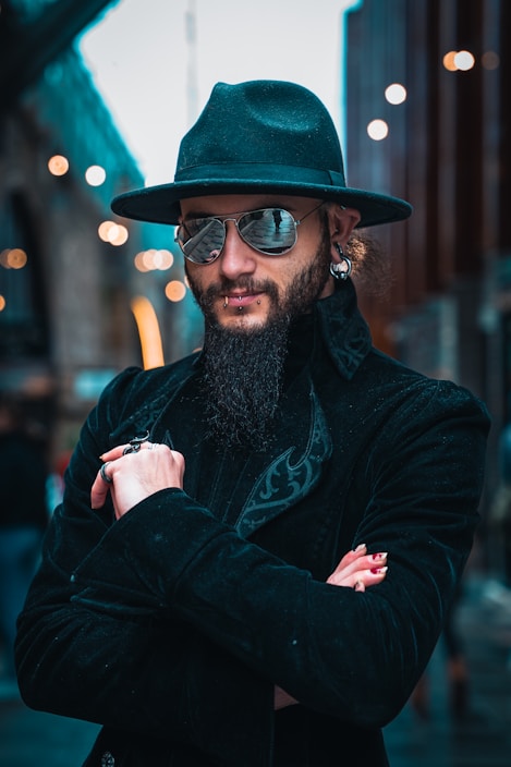 A person with a long beard wearing a black hat and sunglasses, standing confidently with arms crossed. The individual is dressed in a black coat with intricate patterns. The background features a blurred urban setting with scattered lights.