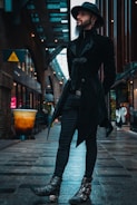 A person dressed in a dark, stylish outfit with a hat stands confidently in an urban alleyway. The cobbled path and architecture convey a modern, city atmosphere. The individual has a long beard and is wearing ornate boots, a tailored coat, and a brimmed hat, exuding a gothic or steampunk fashion sense.