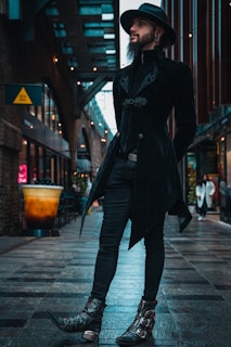 A person dressed in a dark, stylish outfit with a hat stands confidently in an urban alleyway. The cobbled path and architecture convey a modern, city atmosphere. The individual has a long beard and is wearing ornate boots, a tailored coat, and a brimmed hat, exuding a gothic or steampunk fashion sense.