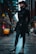 A person dressed in a dark, stylish outfit with a hat stands confidently in an urban alleyway. The cobbled path and architecture convey a modern, city atmosphere. The individual has a long beard and is wearing ornate boots, a tailored coat, and a brimmed hat, exuding a gothic or steampunk fashion sense.