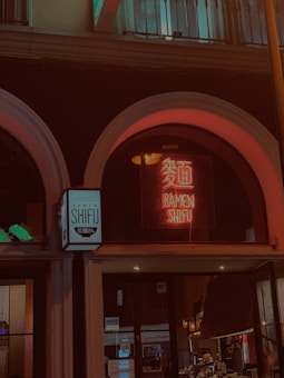 A nighttime street scene featuring a ramen restaurant with a neon sign displaying the words 'Ramen Shifu' both in English and Asian characters. The building is characterized by large arched windows. The atmosphere is dimly lit, giving a warm glow to the surroundings.