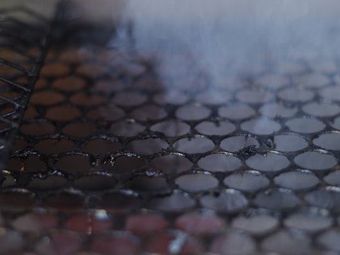 Close-up of a metal grill with circular holes, possibly used for cooking. The surface appears dark and slightly charred, with faint wisps of smoke rising.