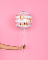 A hand holding a pink and white striped balloon with gold lettering that reads 'Baby Girl', set against a solid pink background. The balloon has decorative gold dots and a ribbon hanging from the bottom.