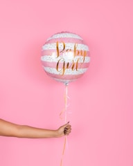 A hand holding a pink and white striped balloon with gold lettering that reads 'Baby Girl', set against a solid pink background. The balloon has decorative gold dots and a ribbon hanging from the bottom.