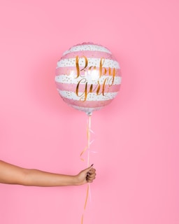 A hand holding a pink and white striped balloon with gold lettering that reads 'Baby Girl', set against a solid pink background. The balloon has decorative gold dots and a ribbon hanging from the bottom.