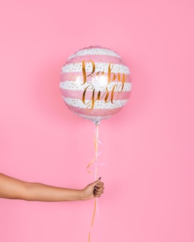 A hand holding a pink and white striped balloon with gold lettering that reads 'Baby Girl', set against a solid pink background. The balloon has decorative gold dots and a ribbon hanging from the bottom.