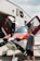 A person stands next to a small, modern single-engine propeller airplane with its doors open. The aircraft is parked in front of a building with large letters, displaying the word 'Aviation.' The plane is primarily red and white, and the person is wearing casual summer clothing, including sunglasses.