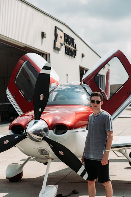 A person stands next to a small, modern single-engine propeller airplane with its doors open. The aircraft is parked in front of a building with large letters, displaying the word 'Aviation.' The plane is primarily red and white, and the person is wearing casual summer clothing, including sunglasses.