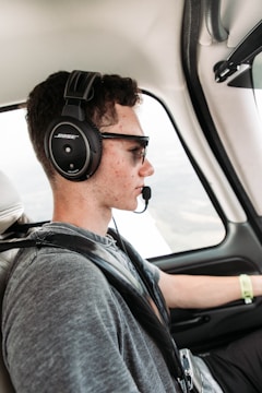 A person is seated in the cockpit of a small aircraft, wearing a gray T-shirt, a black headset with a microphone, and sunglasses. The interior of the plane is visible, including parts of the window and seat. The person appears focused, gazing ahead, possibly piloting the aircraft.