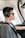 A person is seated in the cockpit of a small aircraft, wearing a gray T-shirt, a black headset with a microphone, and sunglasses. The interior of the plane is visible, including parts of the window and seat. The person appears focused, gazing ahead, possibly piloting the aircraft.