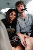 A young woman and a young man are sitting close together, both wearing headsets with microphones, inside a small aircraft. The woman has long dark hair and is smiling, while the man has tousled hair and a beard. They are holding electronic devices, and the interior of the aircraft is visible in the background.