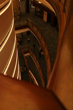 A multi-level indoor structure features modern architecture with curved wooden railings and soft ambient lighting. People can be seen walking on the lower level, and the perspective looks down from an upper floor.