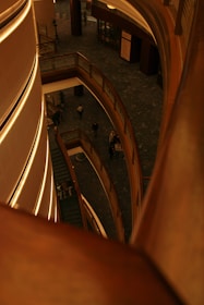 A multi-level indoor structure features modern architecture with curved wooden railings and soft ambient lighting. People can be seen walking on the lower level, and the perspective looks down from an upper floor.