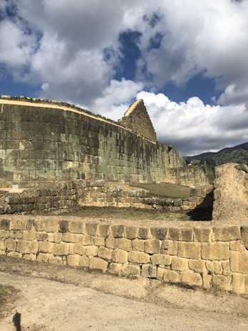 A stone ruin with distinctive ancient architecture, featuring large stone blocks forming walls and a pyramid-like structure. The site appears to be historical, set under a partly cloudy sky with mountains visible in the background.