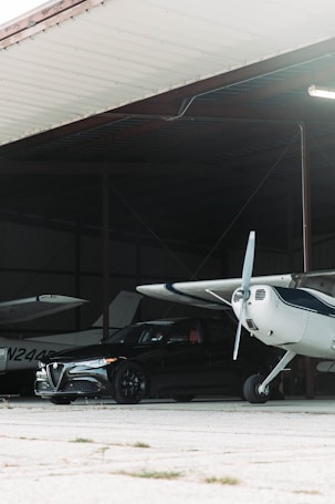 A small private aircraft is parked inside a hangar alongside a sleek black car. The hangar is constructed of metal with an overhead roof, casting shadows within. The aircraft has a white exterior with blue accents and is positioned next to the car. The car, showing a luxury brand symbol on the front, is sleek and polished, contributing to an atmosphere of sophistication.