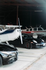 Inside a dimly lit hangar, a collection of vehicles and aircraft are parked. A small plane with a blue stripe stands prominently in the foreground, next to a sleek black sports car and another plane with a red stripe towards the back.