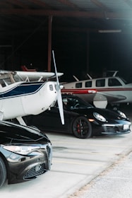 Inside a dimly lit hangar, a collection of vehicles and aircraft are parked. A small plane with a blue stripe stands prominently in the foreground, next to a sleek black sports car and another plane with a red stripe towards the back.