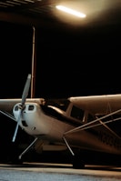 A small single-engine airplane is parked inside a dimly lit hangar. The aircraft is primarily white with black stripes and has a visible propeller at the front. It is partially obscured by shadows, with a beam of artificial light illuminating the area around the plane.