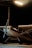 A small single-engine airplane is parked inside a dimly lit hangar. The aircraft is primarily white with black stripes and has a visible propeller at the front. It is partially obscured by shadows, with a beam of artificial light illuminating the area around the plane.