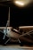 A small single-engine airplane is parked inside a dimly lit hangar. The aircraft is primarily white with black stripes and has a visible propeller at the front. It is partially obscured by shadows, with a beam of artificial light illuminating the area around the plane.