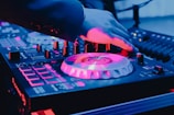 A DJ's hand is adjusting controls on a music mixer, illuminated by vibrant pink, blue, and purple lights. The equipment displays various buttons, sliders, and rotating plates, hinting at a lively, energetic setting.