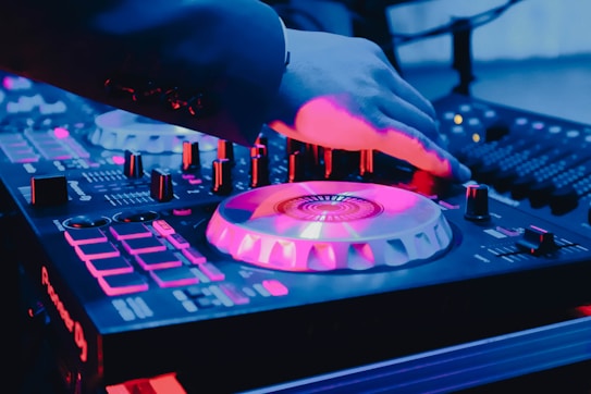 A DJ's hand is adjusting controls on a music mixer, illuminated by vibrant pink, blue, and purple lights. The equipment displays various buttons, sliders, and rotating plates, hinting at a lively, energetic setting.