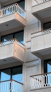 Several balconies of a modern apartment building with white railings and beige outer walls. The balconies have a minimalist design with geometric railing patterns. The glass doors and windows are dark-tinted and reflective.