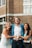 A group of three young people, two women and one man, stand together in front of a brick building, smiling and laughing. The man is in the center wearing a light blue blazer, while the women are on either side, one in a white dress and the other in a blue top. They are making a heart shape with their hands.